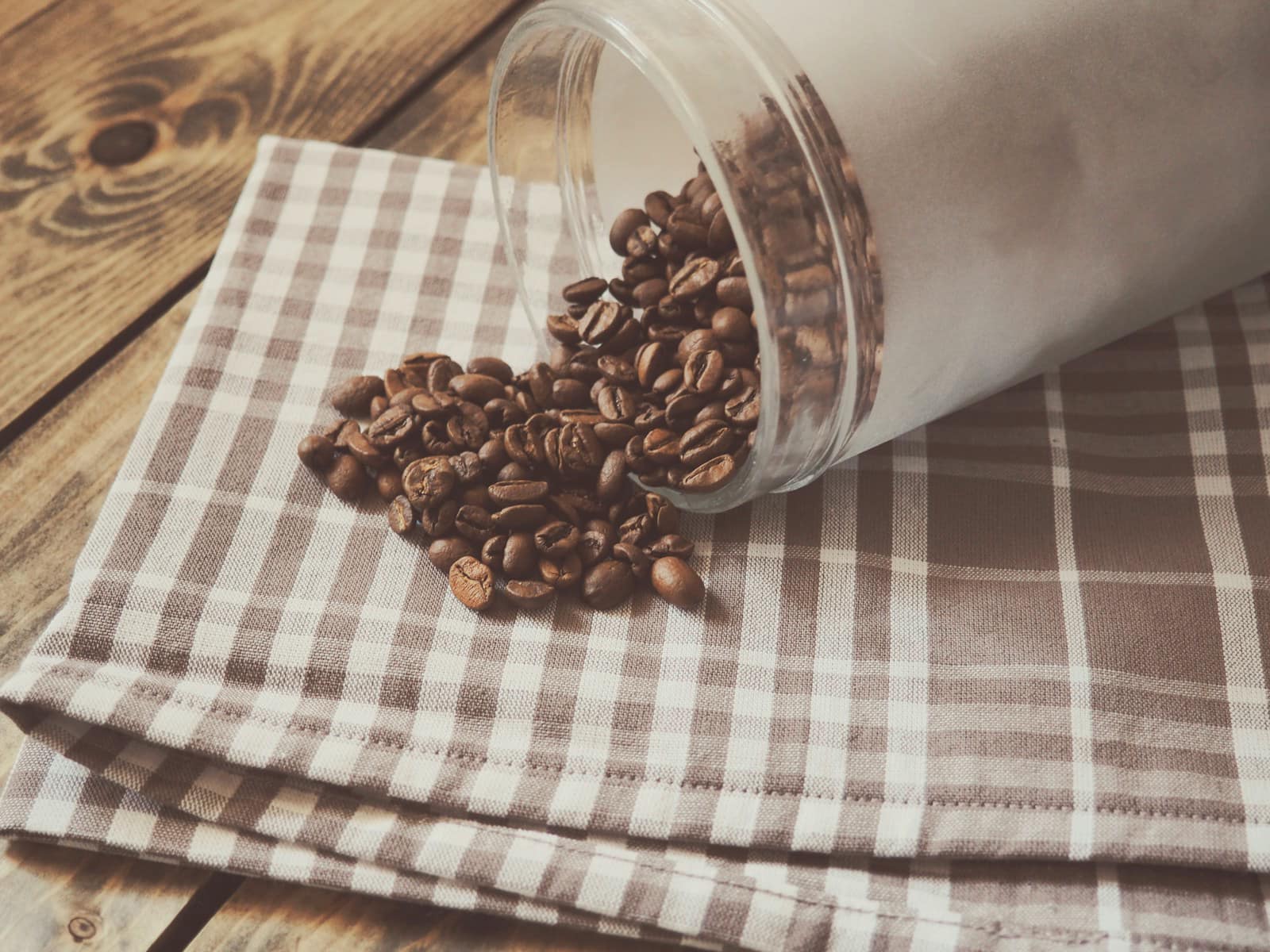 Glass coffee jar tipped on its side with roasted beans spilled onto a checkered cloth